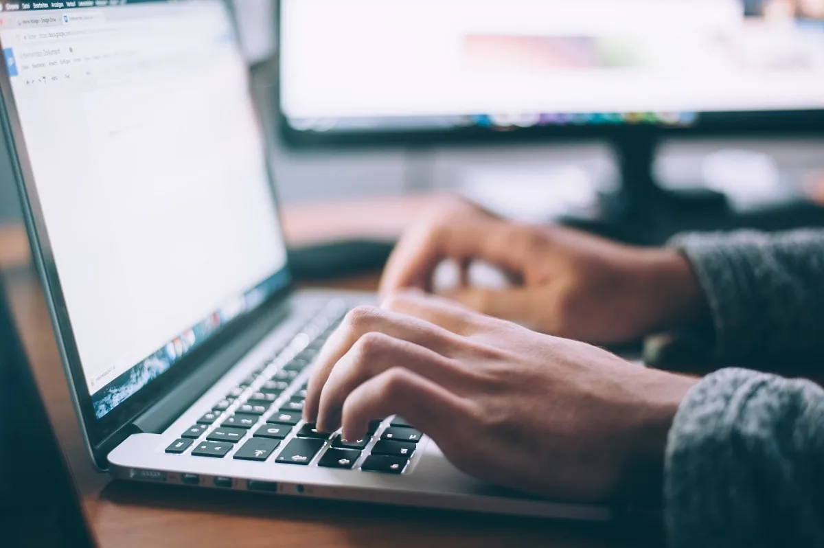 Hände tippen auf einer Laptop-Tastatur, im Hintergrund ein Bildschirm mit geöffneter Website
