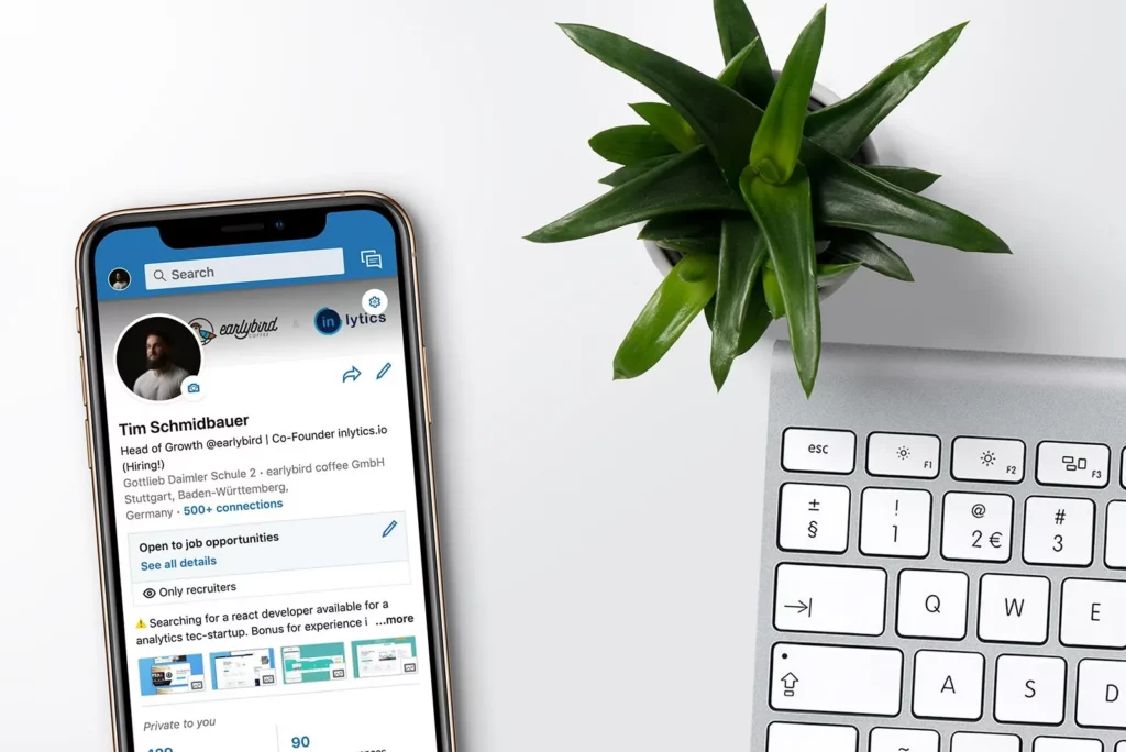 Ein Smartphone mit geöffnetem LinkedIn-Profil, daneben eine Tastatur und eine Zimmerpflanze