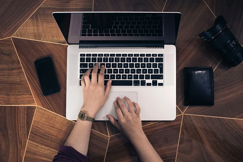 Blick von oben auf einen Holzschreibtisch: Hände an einem Laptop, daneben Smartphone und Kamera