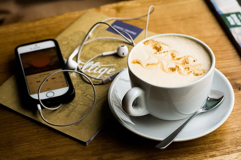 Auf einem Holztisch liegen ein Smartphone mit Kopfhörern und eine Tasse Cappuccino
