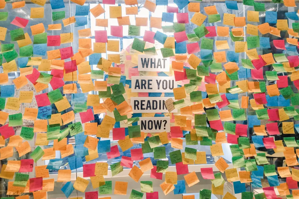 Wand voller bunter Haftnotizen, in der Mitte ein Schild mit der Frage „What are you reading now?“